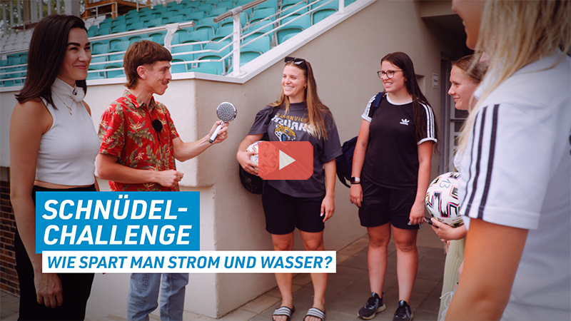 Sarah und Christian auf einem Fußplatz mit der Frauenmannschaft des FC Schweinfurt 05 (Schnüdel). Christian präsentiert einen Sparduschkopf. In der unteren linken Ecke steht: Schnüdelchallenge - wie spart man Strom und Wasser?